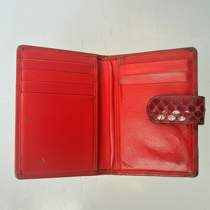 A red leather bifold wallet shown open, featuring a textured python-patterned snap tab closure on the right side, photographed on a light gray reflective surface.
